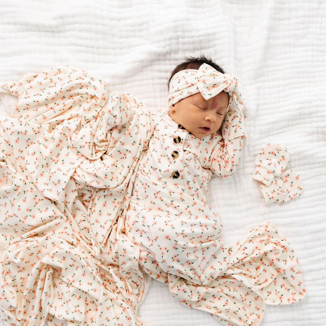 lottie headband bundle newborn girl wearing knotted gown, swaddle blanket, knot headband and mittens laying on bed.